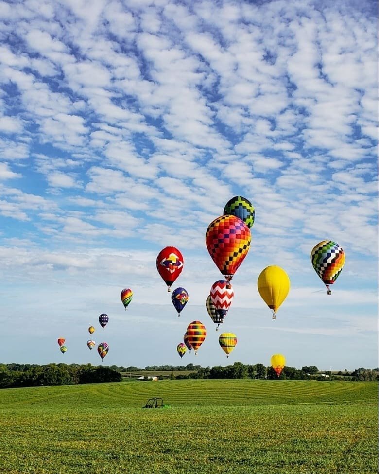 A vibrant array of colorful hot air balloons floats above a lush green field against a backdrop of a partly cloudy blue sky. The scene in Indianola is as lively and captivating as the local real estate, with both offering unique patterns and hues that enchant every observer.