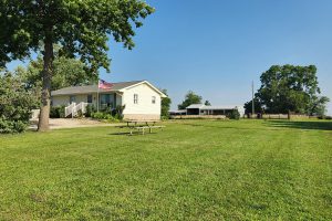 A charming, single-story house with a sloped roof and American flag adorns a vast, green lawn in Indianola's picturesque real estate market. A picnic table graces the foreground, while a barn rests under a clear blue sky, all embraced by surrounding trees.