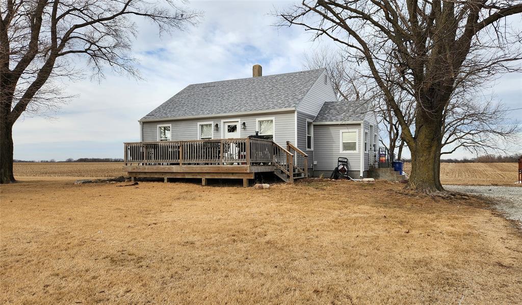 Exterior-1 A small gray house with a gable roof sits surrounded by an expansive field. The house features a wooden deck, bare winter trees, and a gravel path leading to it. The sky is overcast with hints of blue.