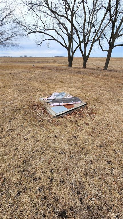 Exterior-10 A broken metal sign lies on dry grass surrounded by fallen leaves. In the background, bare trees stand against a cloudy sky, and an open field stretches into the distance.