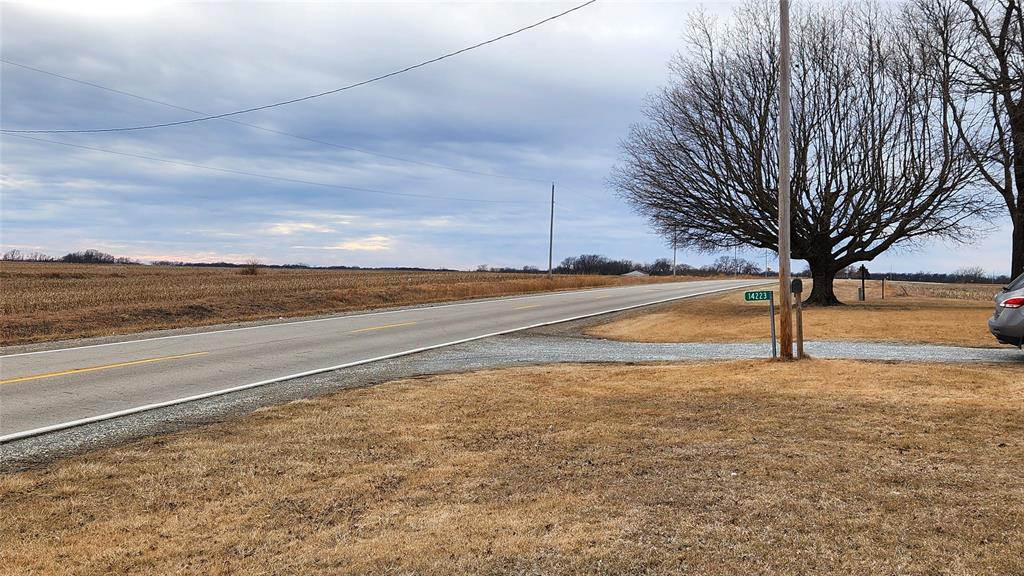 Exterior-11 A rural intersection with a paved road and an empty field under a cloudy sky. A bare tree stands on the right, next to a parked car partially visible. A street sign is nearby, and power lines run along the road.