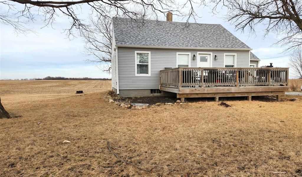 Exterior-12 Small gray house with a shingled roof and a wooden deck on the side, situated on a barren lawn. Sparse trees surround the area, and vast, empty fields stretch into the distance under a cloudy sky.