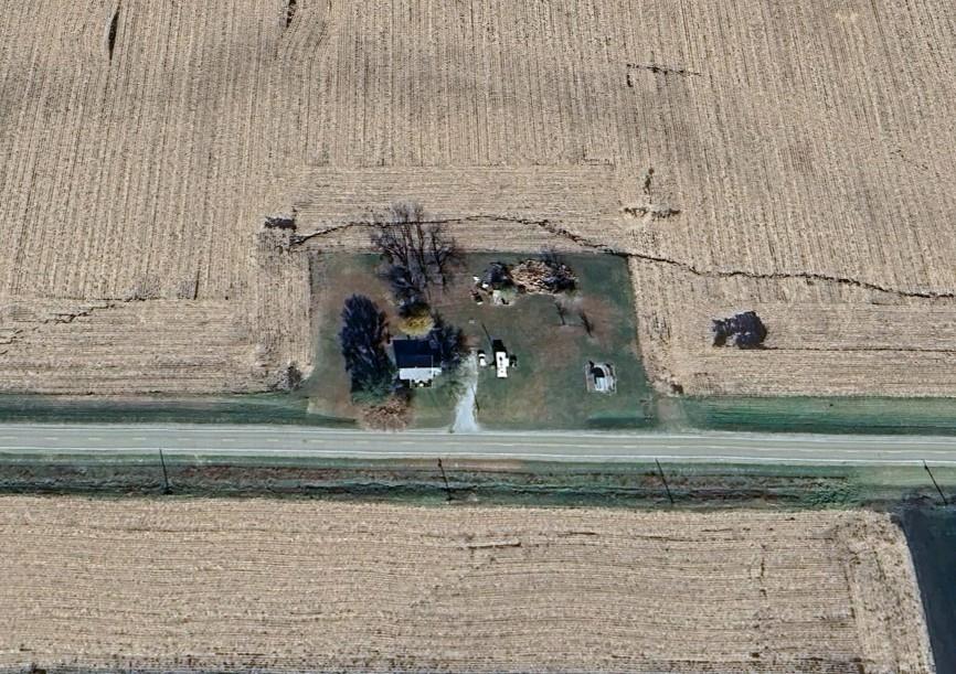 Exterior-13 Aerial view of a rural landscape with a small house and two vehicles on a grassy plot, surrounded by large expanses of tilled farmland. A straight road runs horizontally across the foreground. Sparse trees are scattered around the property.