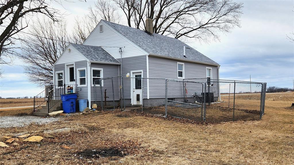 Exterior-2 A small, gray house with a gabled roof is surrounded by a metal chain-link fence. There are leafless trees behind it and a trash bin near the steps. The yard is brown and dry, and the sky is overcast.