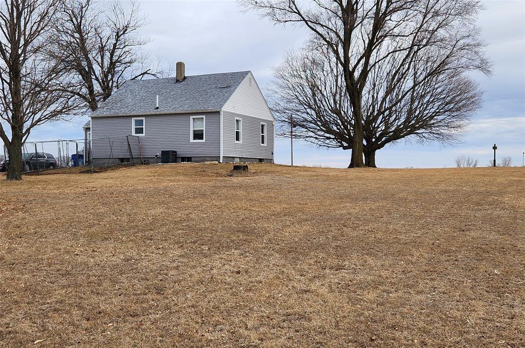 Exterior-3 A small gray house with a sloped roof stands on a grassy field. Leafless trees border the house, and a cloudy blue sky is visible in the background. The ground is covered with dry grass, with no other structures nearby.