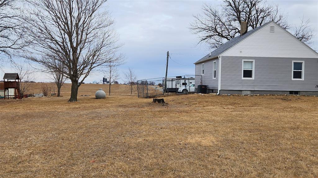 Exterior-4 A gray house with a gable roof stands on a grassy yard. A bare tree and a large spherical stone are in the foreground. A playset is visible to the left, and a trailer is parked next to the house. Overcast sky in the background.