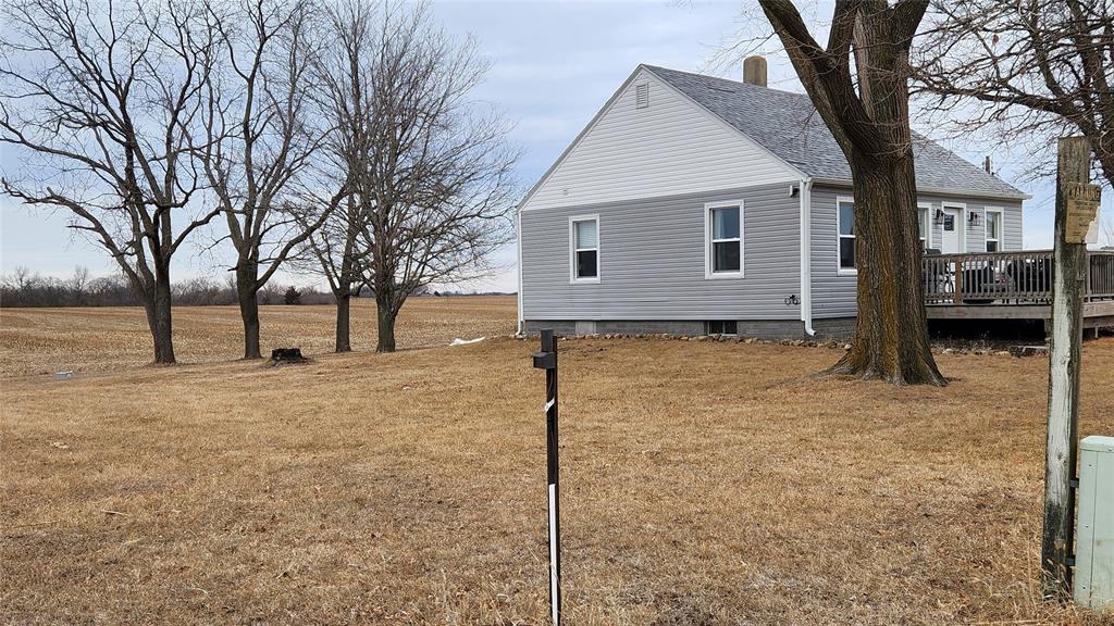 Exterior-5 A small gray house with a sloped roof sits next to leafless trees on a dry, grassy lawn. A field stretches into the distance under an overcast sky. A post is visible in the foreground.
