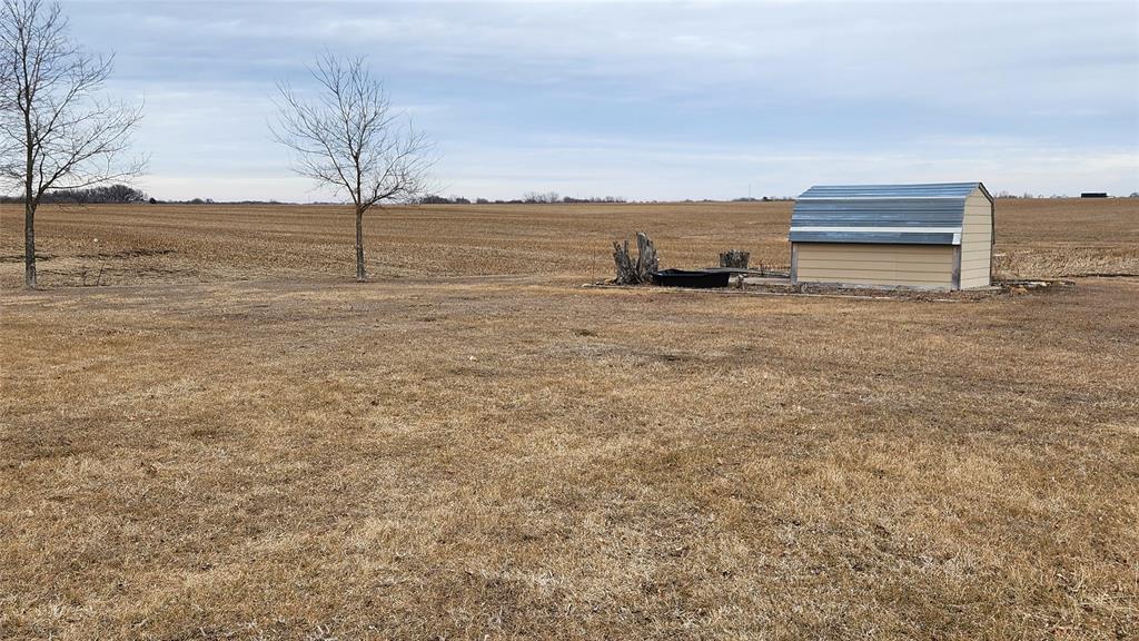 Exterior-7 A vast field with dry, brown grass under a cloudy sky. There are two bare trees in the foreground and a small structure with a metal roof on the right side, next to some rocks. Farmland extends into the distance.