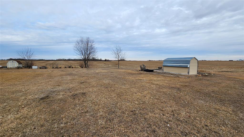 Exterior-8 A vast field with dry grass and a cloudy sky. In the foreground is a modern storage shed with a curved metal roof. Two leafless trees stand nearby. The flat landscape extends into the distance.