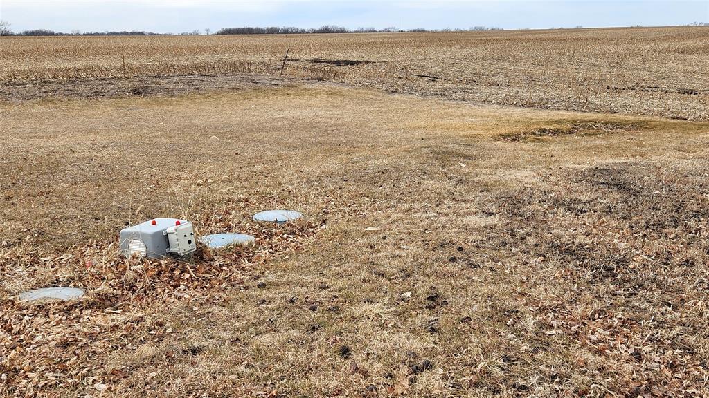 Exterior-9 A vast, flat field with patches of dry grass and scattered leaves. Three white utility covers are visible on the ground, with one surrounded by leaves. The sky is overcast, creating a subdued atmosphere in the open landscape.