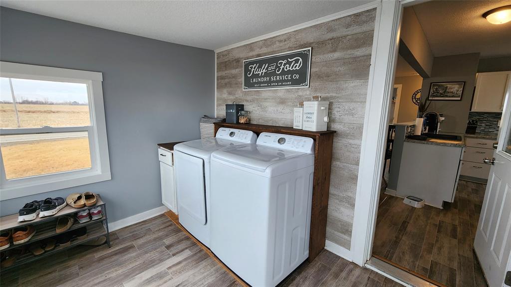 Interior-10 Laundry room with a washing machine and dryer against a wood-paneled wall. Shelves hold detergent and decor. A small window shows an outdoor view, and several pairs of shoes are on a rack. The room connects to a kitchen with wood flooring.