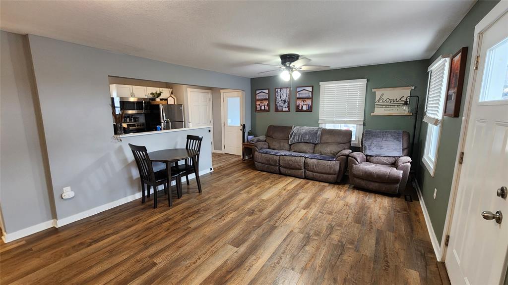 Interior-2 Living room with wooden floors, gray walls, and a ceiling fan. Features a cozy gray sofa, a small black dining table with two chairs, wall art, and a kitchen view through an open counter. Windows allow natural light.
