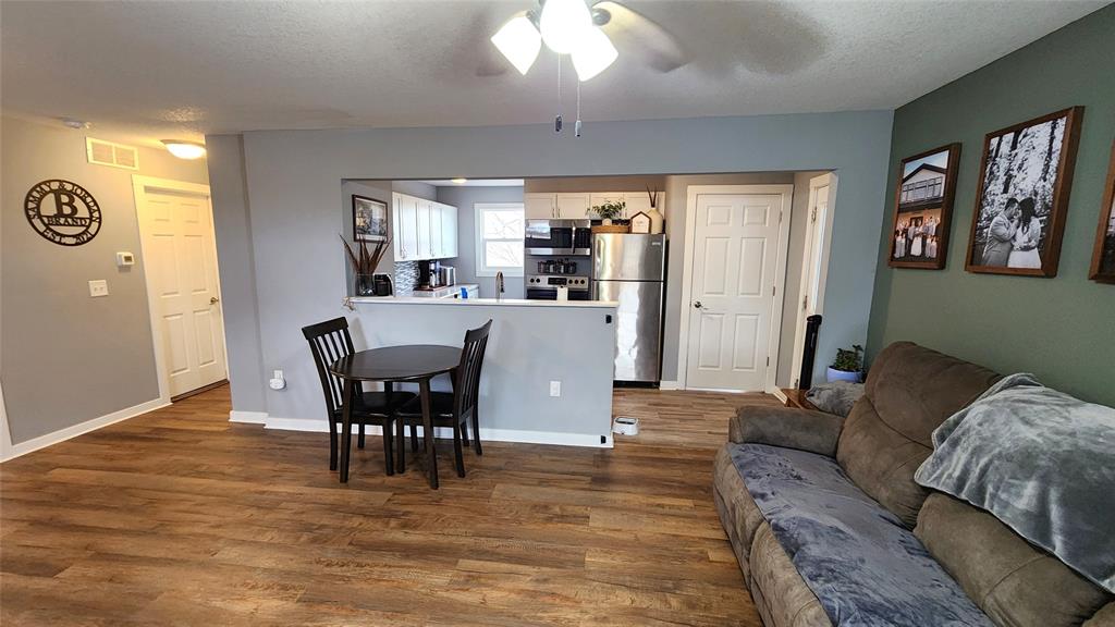 Interior-3 Modern living room with wooden flooring, a gray couch, and a small black dining table with two chairs. The kitchen has stainless steel appliances and white cabinets. Walls are painted in gray and green tones, with framed photos hanging on one wall.