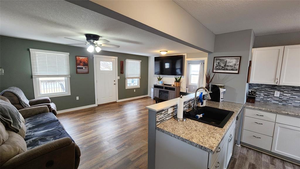 Interior-4 Open-plan living room and kitchen with wooden floors. The living area features a ceiling fan, brown sofa, large TV on a wall mount, and a small fireplace. The kitchen has white cabinets, granite countertops, and a tile backsplash.