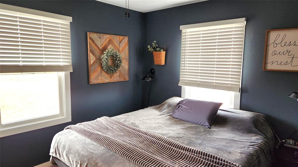 Interior-5 A cozy bedroom with dark blue walls features a bed with gray bedding and a striped throw blanket. Two windows with blinds brighten the room. Decorations include a wooden framed wreath and a sign reading "bless our nest.