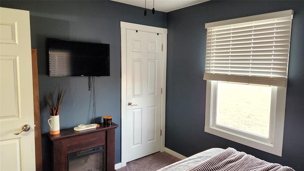 Interior-6 A cozy bedroom with dark blue walls features a wall-mounted TV next to a white door. A window with blinds partially open lets in light. Below the TV is a small electric fireplace with decor, and a neatly made bed is in the lower right corner.