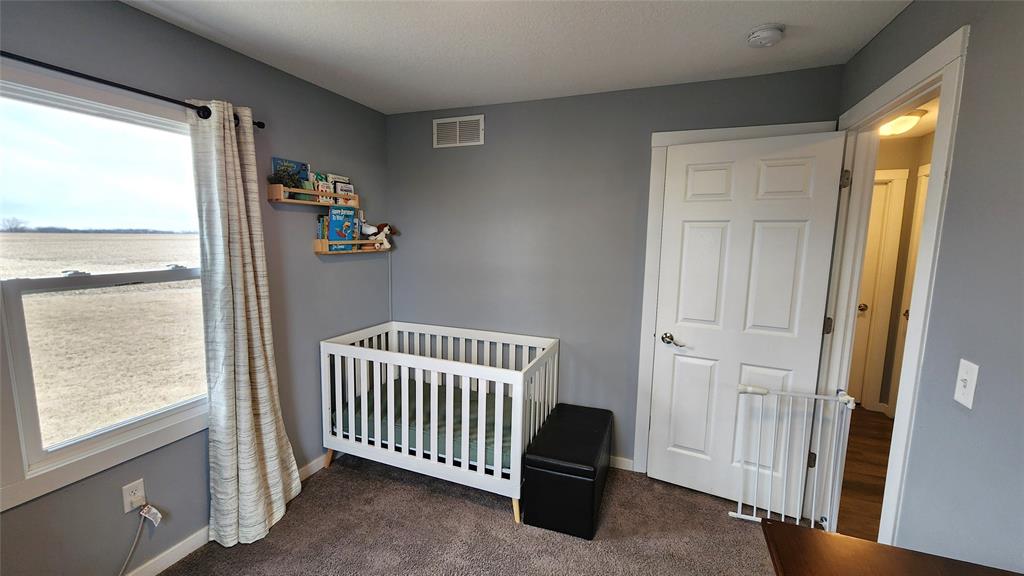 Interior-9 A nursery room with gray walls features a white crib, a small black ottoman, and a white safety gate by the door. A window with light curtains shows a view of a field. Shelves above the crib display books and a stuffed animal.