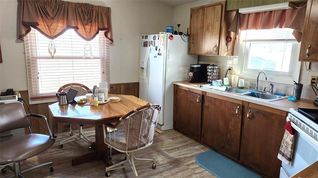 A cozy kitchen with wooden cabinets, a white fridge adorned with magnets, a window above the sink, blinds with tan curtains, and a round wooden table surrounded by chairs. The floor is light wood, and the space has a vintage feel.