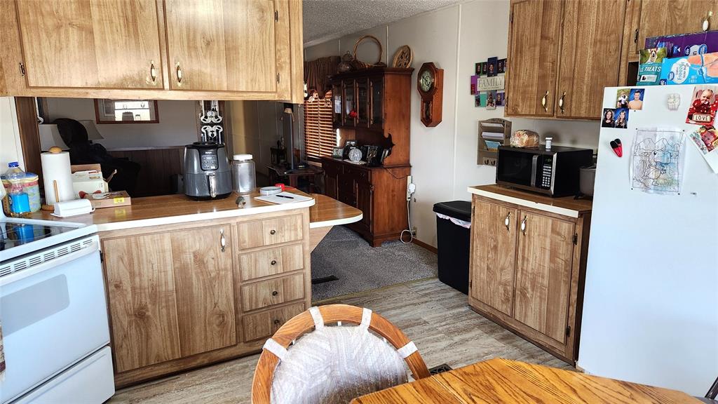 A cozy kitchen with wood cabinets and beige countertops. A white refrigerator with magnets and a microwave sit against the right wall. A wooden chair and dining table are in the foreground. A narrow hallway leads to a living area in the background.