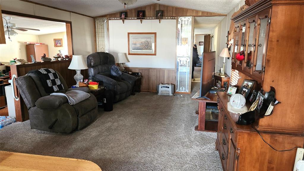 A cozy living room with a reclining sofa and various household items scattered around. A wooden cabinet with decorative items and a TV is on the right. The walls are paneled, and there is carpet on the floor. A fan can be seen on the ceiling.