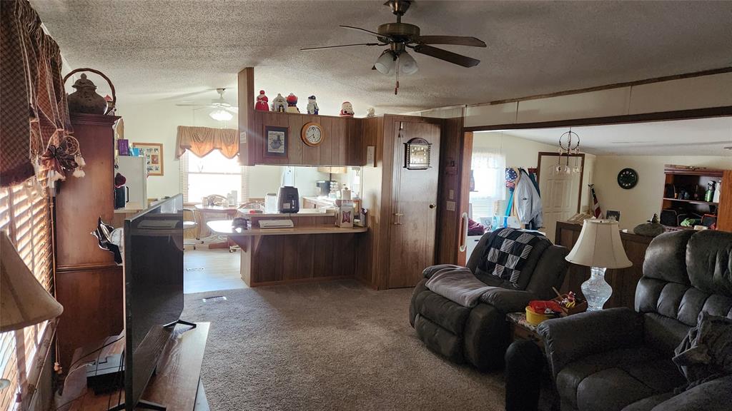 A cozy living room with brown carpeting features a ceiling fan, plush armchairs, and a TV. The room is adorned with knick-knacks and a lamp. A wooden partition divides it from the kitchen, visible in the background with a dining area.