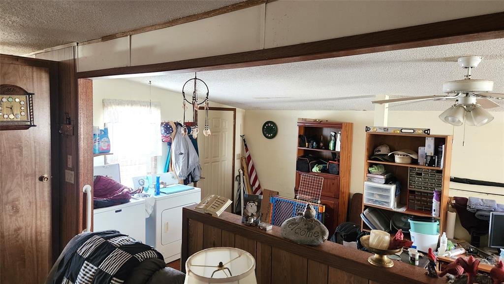 A cluttered room with a laundry area featuring a washer and dryer. The room has wooden furniture, a ceiling fan, and various items scattered around, including a clock, an American flag, and storage shelves filled with miscellaneous objects.
