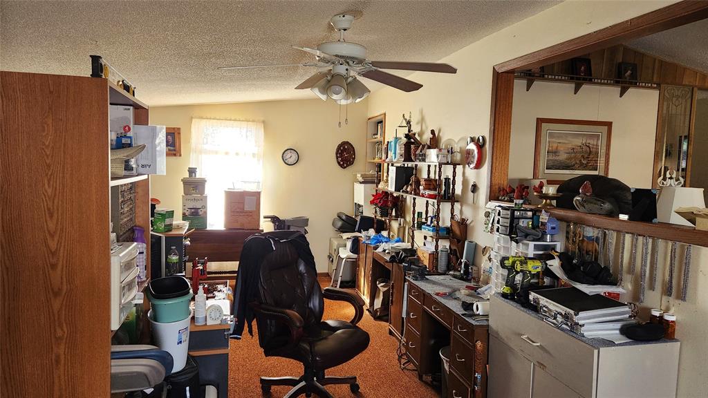 Cluttered home office with an office chair, shelves full of various items, and a desk with tools and papers. A ceiling fan hangs above, and there’s a window with light filtering in, along with a wall clock and framed art.