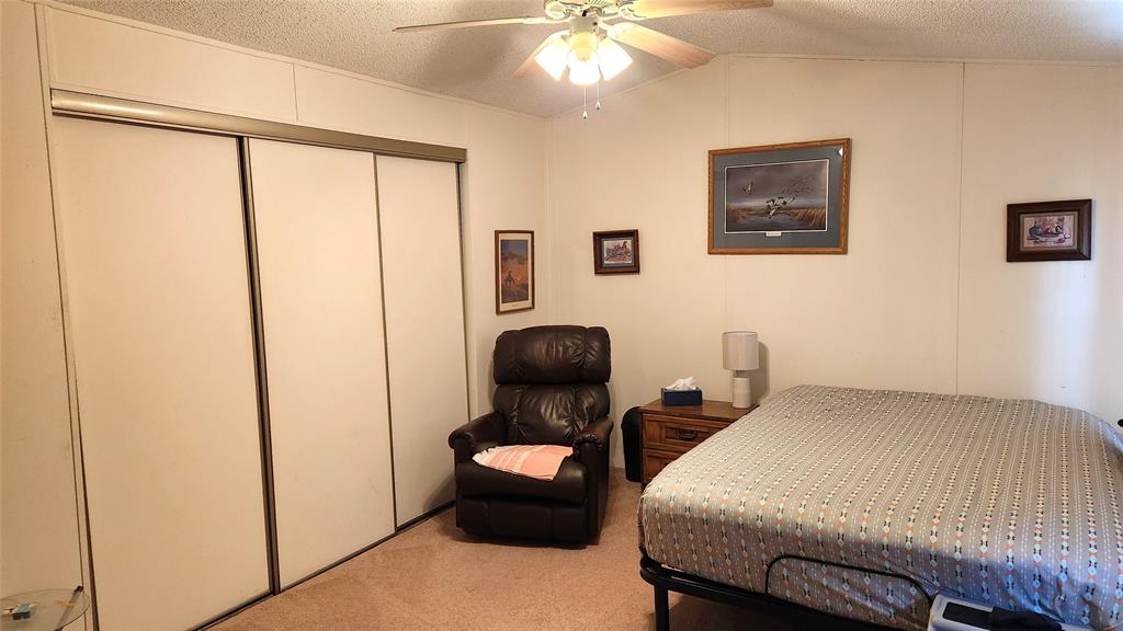 A bedroom with a bed, patterned bedding, brown recliner, and small wooden nightstand with a lamp. A ceiling fan with lights is on. Three framed pictures hang on the walls, and a sliding closet door is to the left.