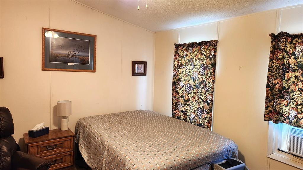 A bedroom with cream-colored walls, featuring a bed with a patterned gray comforter, a wooden nightstand with a lamp and a tissue box, and two windows with floral curtains. Framed artwork decorates the walls. An air conditioner is below one window.