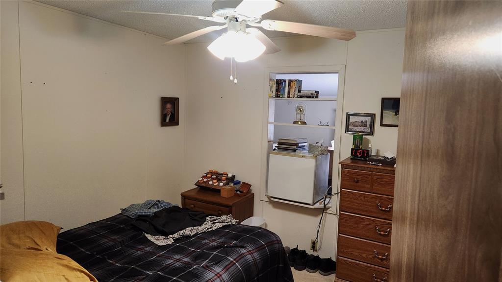 A small bedroom with a ceiling fan and a checkered bedspread on an unmade bed. A dresser with several items on top stands beside the bed. The walls have framed pictures, and a doorway reveals a lit adjacent room with books and a lamp on shelves.
