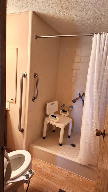 Accessible bathroom with a shower. The shower has a curtain, grab bars, and a white shower chair with back support. Wood flooring is visible in front of the shower, and part of a toilet is seen to the left.