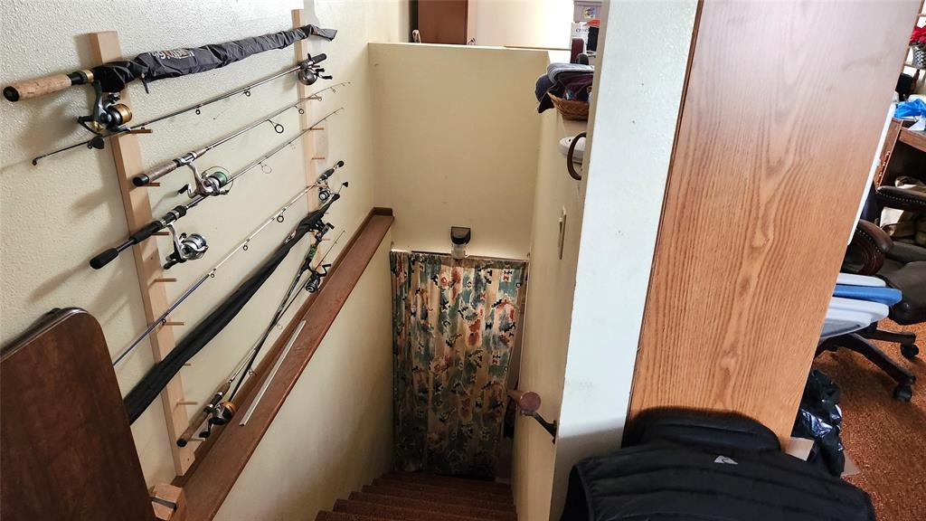 A staircase leads down to a closed curtain featuring a nature pattern. Fishing rods are mounted on a wall rack to the left. Office chairs and various items are partially visible on the right.