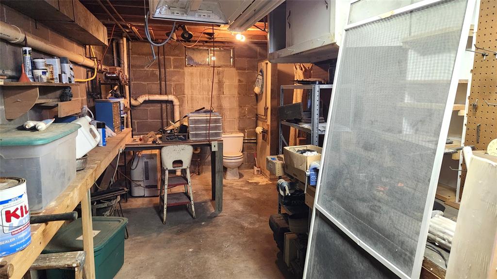 A cluttered basement workshop with shelves holding various tools and storage bins. A table with tools is in the center, and a toilet is visible in the background. Exposed pipes run along the ceiling and walls.