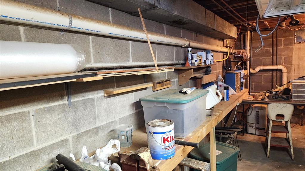 A cluttered basement workshop with concrete walls. A wooden workbench is filled with paint cans, a plastic storage container, and various tools. Shelves above hold more items. Pipes run along the wall, and an electrical box is visible.