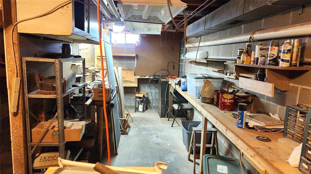 A cluttered workshop with shelves and workbenches lining the walls. Various tools, paints, and materials are scattered around. A coffee can and cleaning supplies are visible. The space is lit by a window and overhead lights.