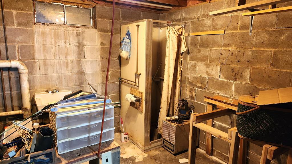A cluttered basement with concrete walls contains a shower, a small sink, and various miscellaneous items, including a plastic chest of drawers, a cardboard box, and a rolled-up curtain. A small window is located on the upper left wall.