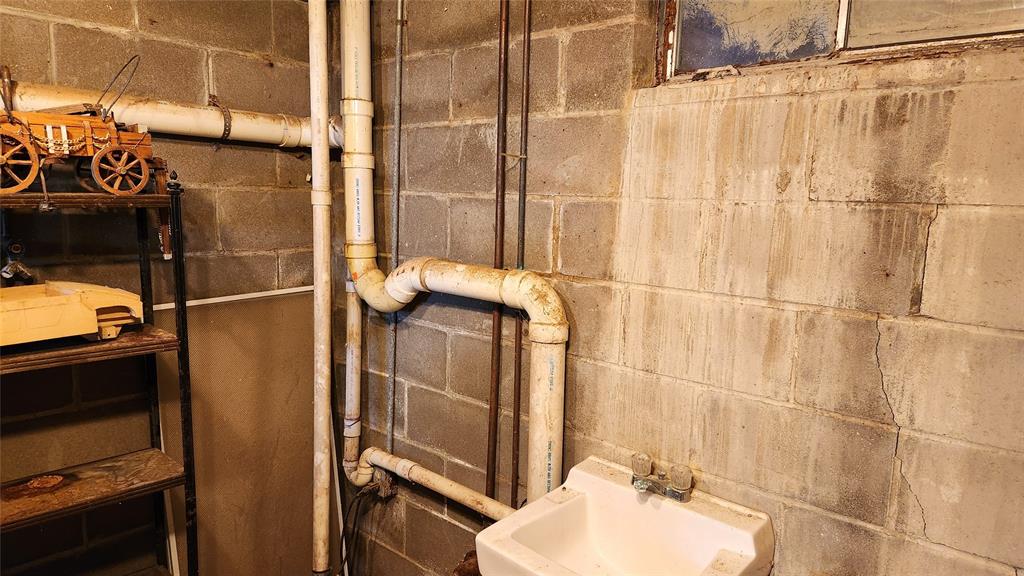 A small basement or utility room with exposed pipes running along a gray cinder block wall. A white sink is installed beneath a window with peeling paint. On a metal shelf, there's a small model wooden cart and assorted items.