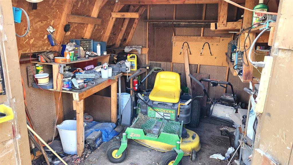 Inside a cluttered wooden shed, there's a green and yellow lawn mower at the center. Shelves on the left hold various tools, cans, and containers. The shed has a rustic appearance with exposed wooden beams and tools hanging on a pegboard.