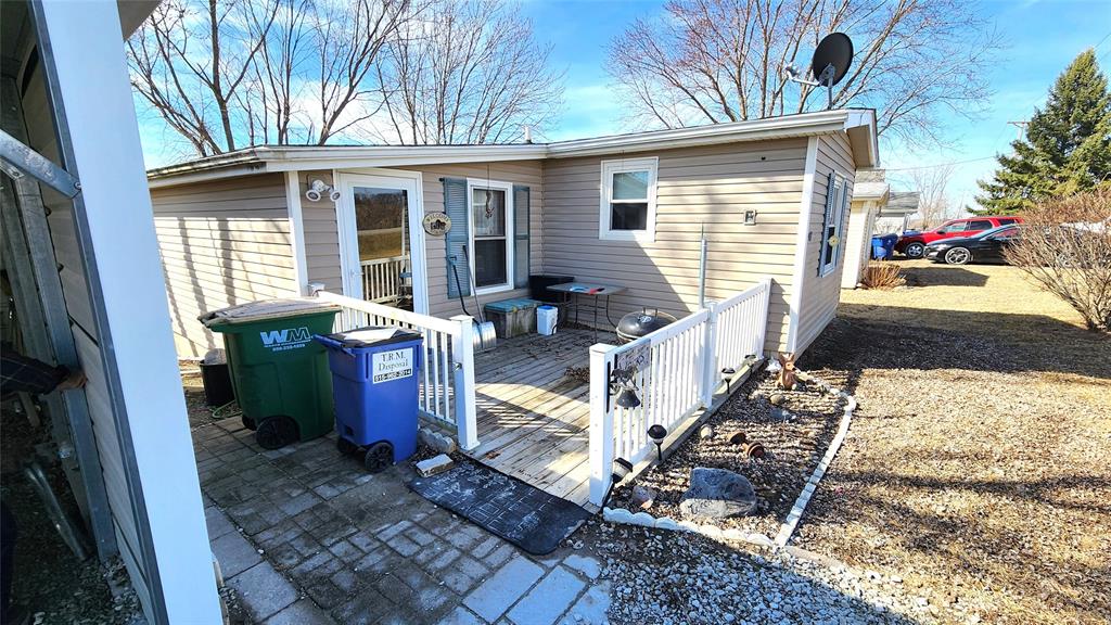 A small beige house with a white railing along a wooden ramp leading to the entrance. There are blue and green recycling bins on the deck. Trees without leaves are in the background under a clear blue sky. A satellite dish is on the roof.
