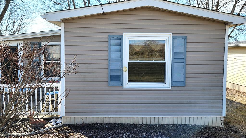 A small, single-story house with beige siding and blue shutters around a window. A porch is partially visible on the left, along with a leafless shrub. The ground is covered with gravel and stones. There are trees and a clear blue sky in the background.