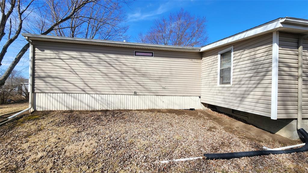 The image shows the exterior side of a beige house with horizontal siding. There are no windows on the wall except for a small one near the roofline. Leafless trees are visible, and a section of the ground is covered in dry grass and pebbles.