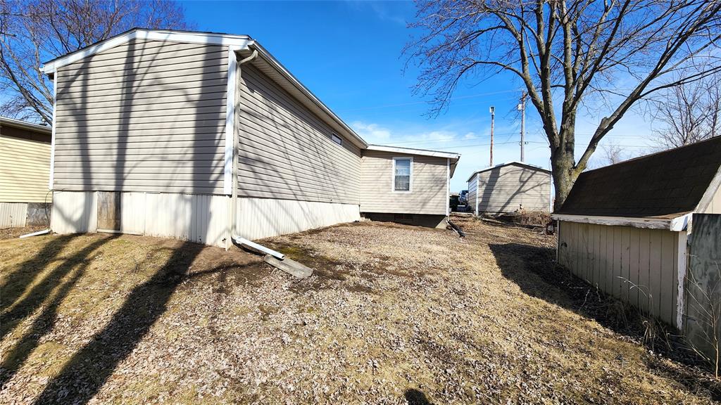 A beige mobile home is positioned on a patchy grassy yard with a nearby tree and a small shed to the right. Shadows of branches are cast on the ground. The sky is clear and blue.