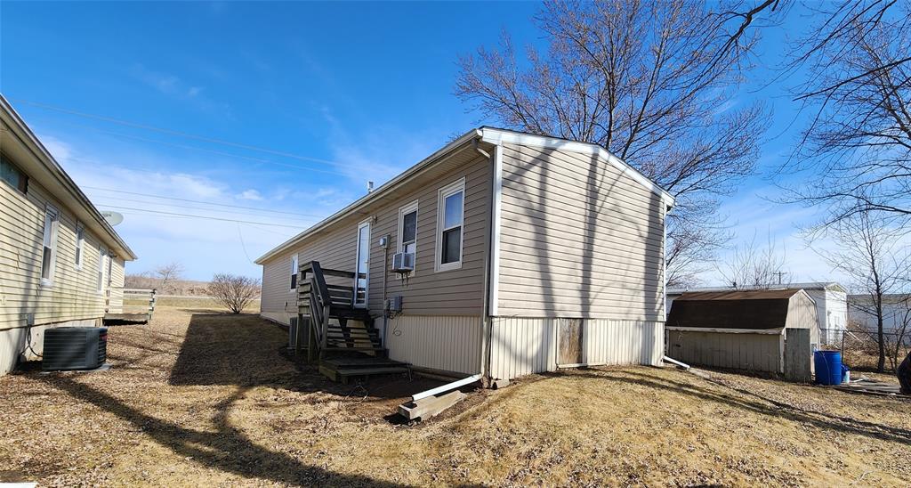 A single-story, beige mobile home with a small set of stairs leading to the entrance. The home sits on a slight hill with dried grass around. Bare trees are visible in the background under a clear blue sky.