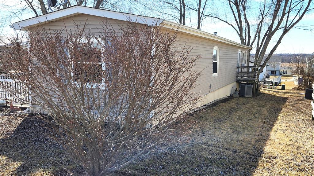 Side view of a beige mobile home with a leafless shrub in the foreground. The home has a small deck with railing and is surrounded by a grassy yard. Bare trees and other houses are visible in the background under a clear blue sky.
