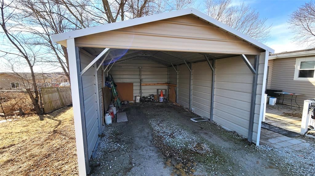 An empty outdoor metal carport with a beige roof and walls, open at the front. Inside, various gardening tools and supplies are stored against the sides. The ground is a mix of gravel and dirt, with dry grass surrounding the structure.
