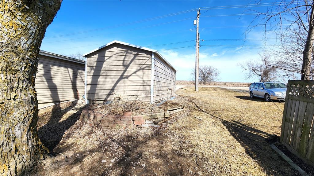 A small shed with gray siding stands near a large tree on a grassy lawn. A brick-bordered garden bed is in the foreground, and a blue car is parked near a wooden fence in the background under a clear blue sky.