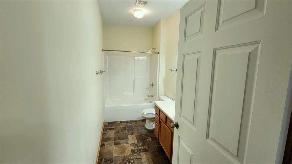 A narrow bathroom with beige walls and a brown tile floor. It features a white bathtub with a shower, a toilet, and a wooden vanity with a sink. The door is partially open, and there's a light on the ceiling.