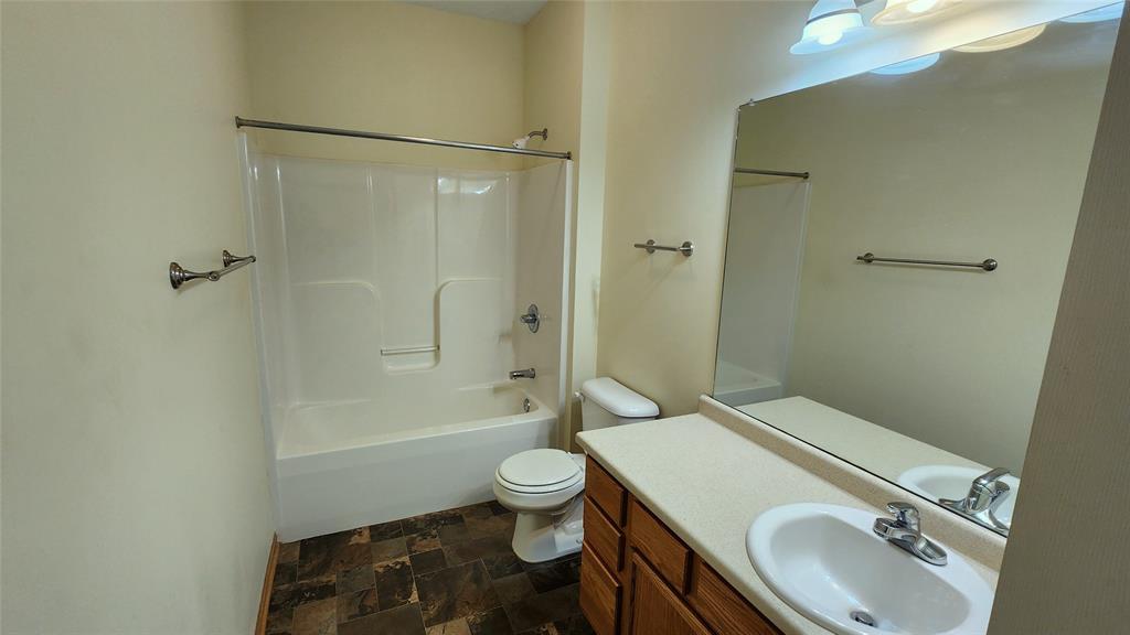 A bathroom with a bathtub-shower combo, a toilet, and a sink with a countertop. There's a large mirror above the sink. The floor is tiled and the walls are light-colored. Towel racks are mounted on the walls.