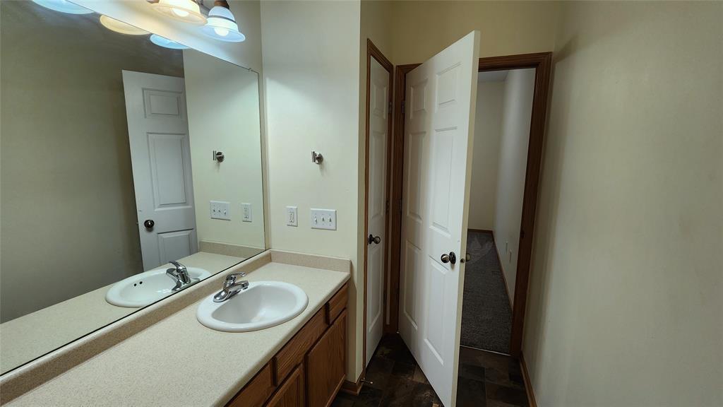 A bathroom with a single sink and beige countertop. There's a large mirror above the sink and light fixtures hanging over it. The walls are off-white, and a partially open door leads to a carpeted hallway.