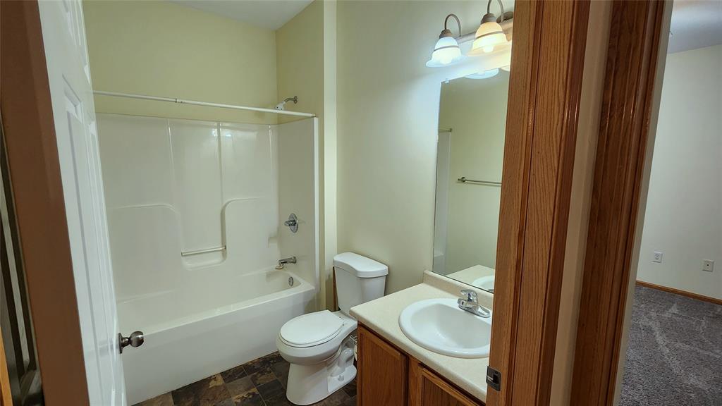 A bathroom with a white bathtub-shower combo, a toilet, and a sink with a wooden cabinet. Above the sink is a mirror and a triple light fixture. The floor is tiled, and the walls are painted light yellow.